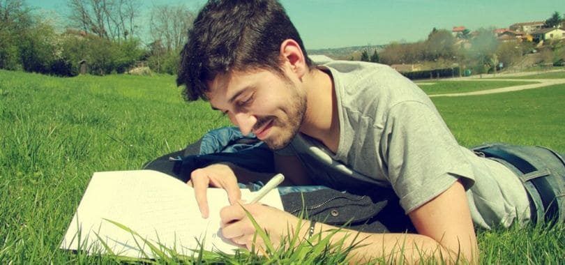 A student lying on a lawn writing down notes on a notebook.