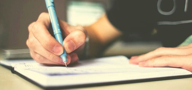 A person holding a blue pencil writing in a notebook.