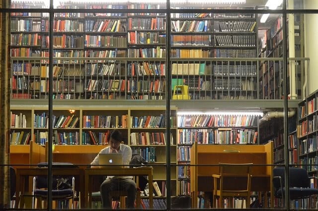 Man studying alone on his Macbook at the library showing bunch of books behind him.