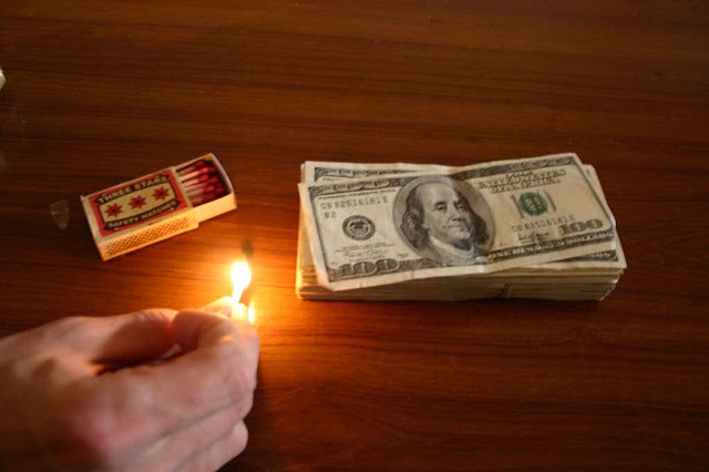 A hand lighting up a match beside a pile of a dollar bill.