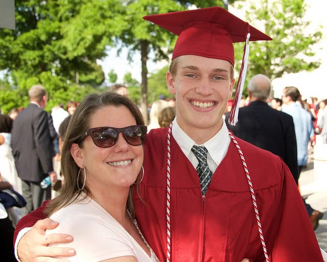A portrait of a young man wearing a red graduation gown with his mom on his side.
