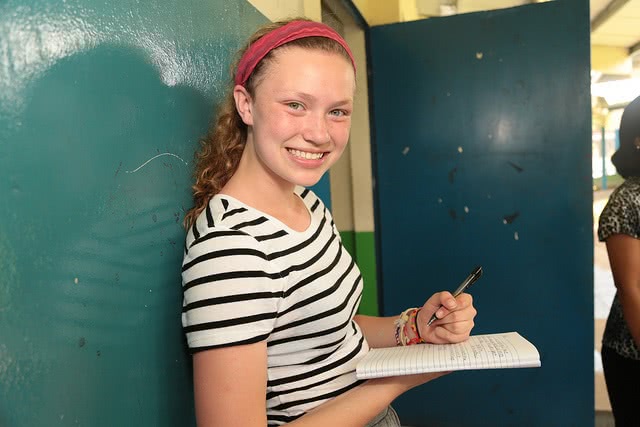 A girl smiling while holding a ballpen and notebook with both hands.