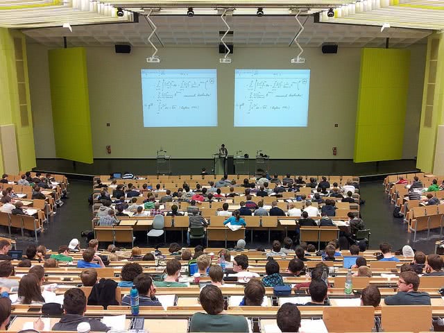 Students sitting in a huge university classroom seen from behind.