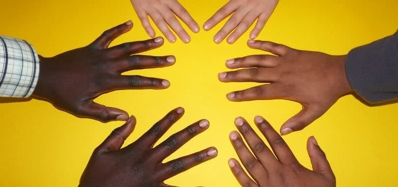 Three people of various races place their hands on a yellow table.